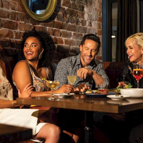 A group of people sit around a table in a cozy, dimly lit room, sharing drinks and conversation, with murals in the background, smiling warmly.