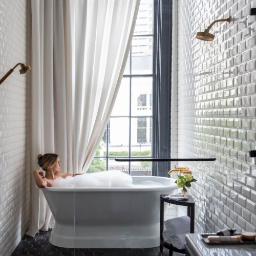 A cozy, sunlit bedroom with a large window, a bed or daybed, a radiator, plants, and soft curtains.