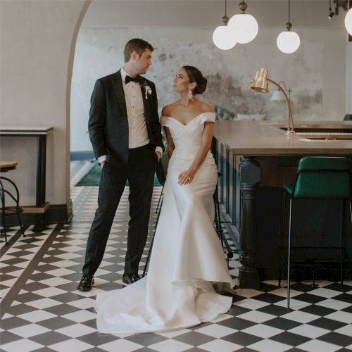 A bride in a white wedding gown and a groom in a black tuxedo stand together, smiling in a stylish black-and-white tiled venue with modern lighting.