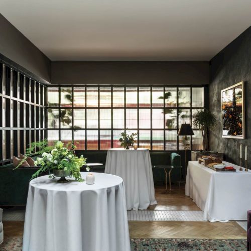 A stylish lounge/boardroom setup with round tables covered in white linens, plants as centerpieces, and a large windowed wall admitting natural light, ending the sentence.