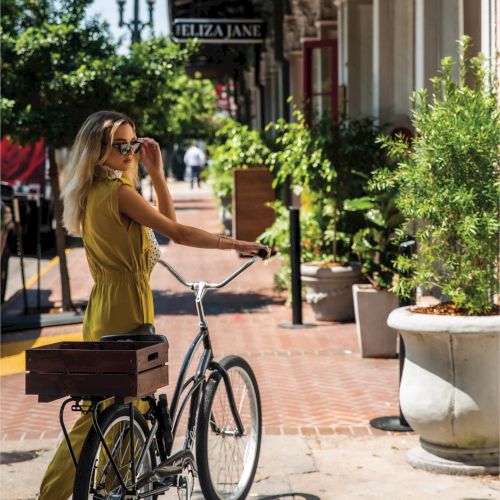 A man and a woman ride bicycles along a sunny street lined with parked scooters and potted plants, chatting beside a cafe.