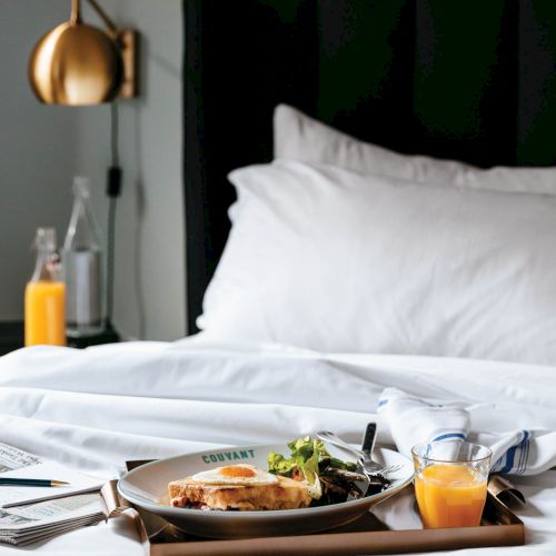 A neat breakfast on a bedside tray: a toasted sandwich, orange juice, coffee, yogurt, and fruit beside a white pillow&mdash;cozy hotel vibes.