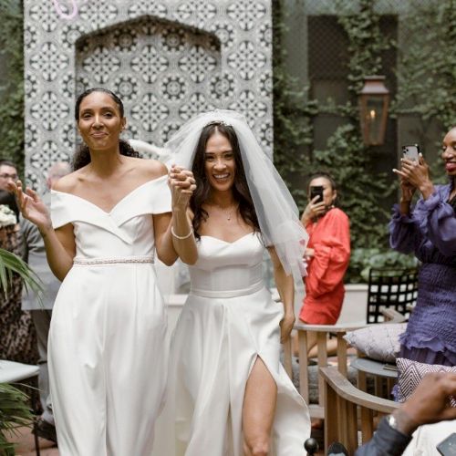 Two brides walk arm in arm, smiling in white gowns at an outdoor wedding; guests cheer and capture the moment.