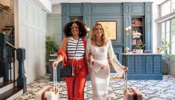 Two stylish women with suitcases stand in a chic hotel lobby, smiling at the camera as they walk in together.
