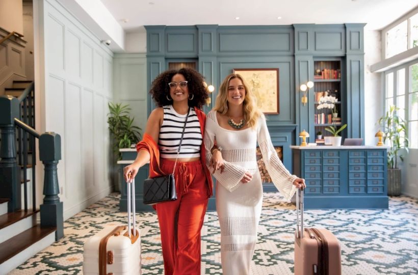 Two stylish women with suitcases stand in a chic hotel lobby, smiling at the camera as they walk in together.