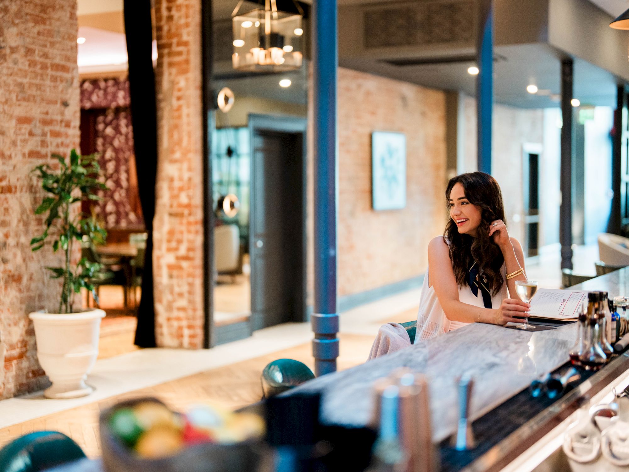 A woman in a light dress sits at a bar counter, leaning on her elbow and gazing to the side in a stylish, brick-walled lounge.