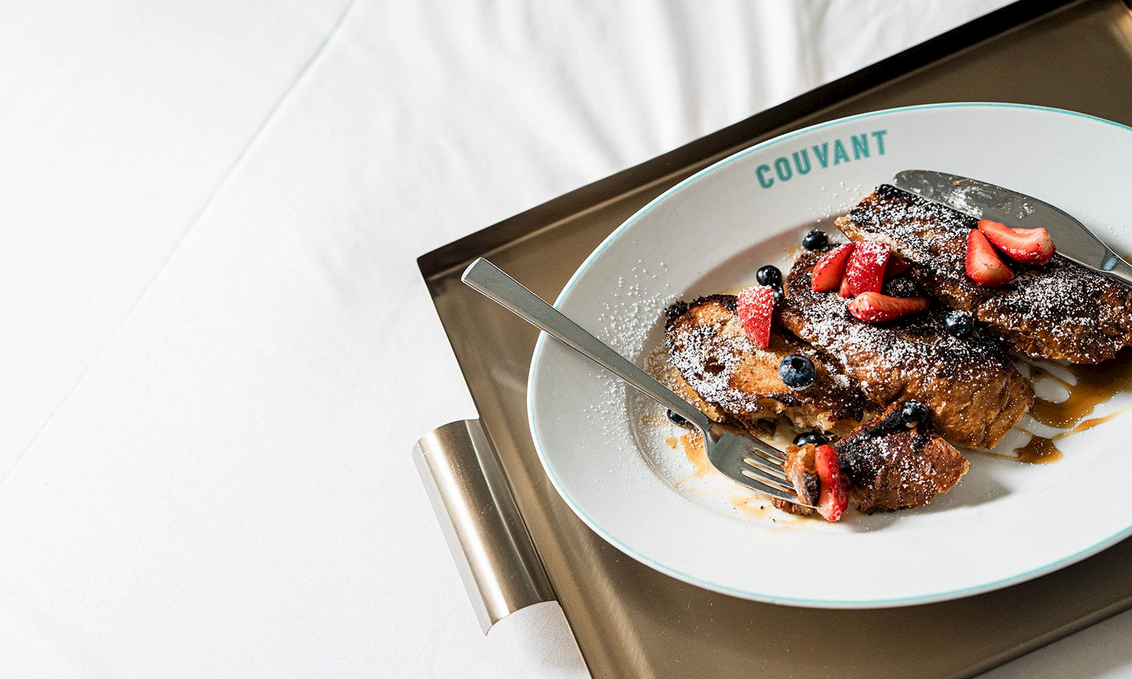 A plate with French toast or pastry topped with strawberries and dusted with powdered sugar sits on a tray; elegant breakfast scene.